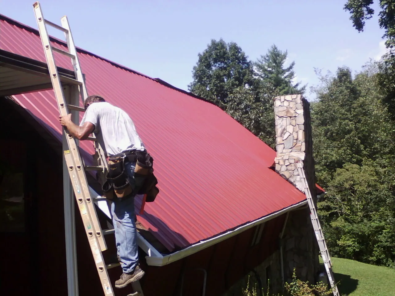 Roofer on a ladder beside a red metal roof and stone chimney — Soffit & Fascia Repair by Roofing Brothers in Powder Springs